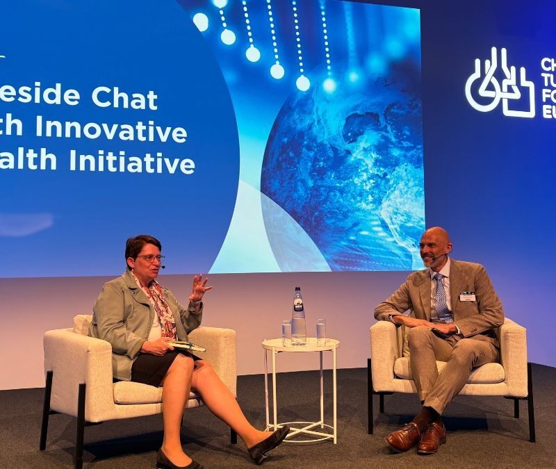 Two people seated on stage in discussion during the "Fireside Chat with Innovative Health Initiative" at a Children's Tumor Foundation Europe event. A screen behind them displays the event title.